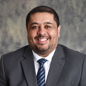  Jose Saavedra-Torres smiles in a studio portrait against a neutral background. He is wearing a gray suit jacket, white dress shirt, and a blue striped tie.
