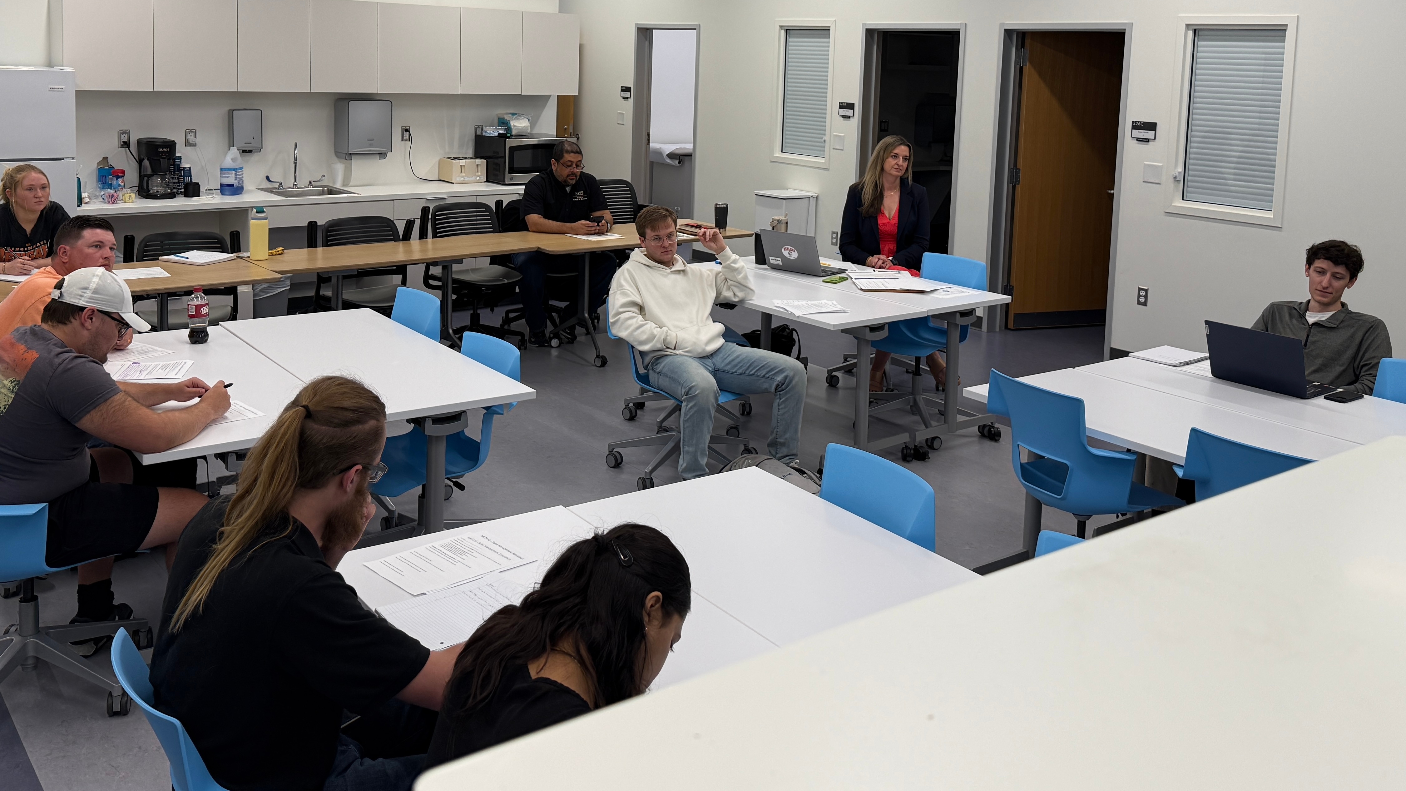 Students and an instructor sit around tables in a modern classroom, engaged in a discussion or workshop. Several students take notes on printed papers while others listen or use laptops. The room includes white tables, blue chairs, and a kitchenette area in the back with cabinets, a sink, and small appliances. The atmosphere suggests a collaborative, hands-on learning session.