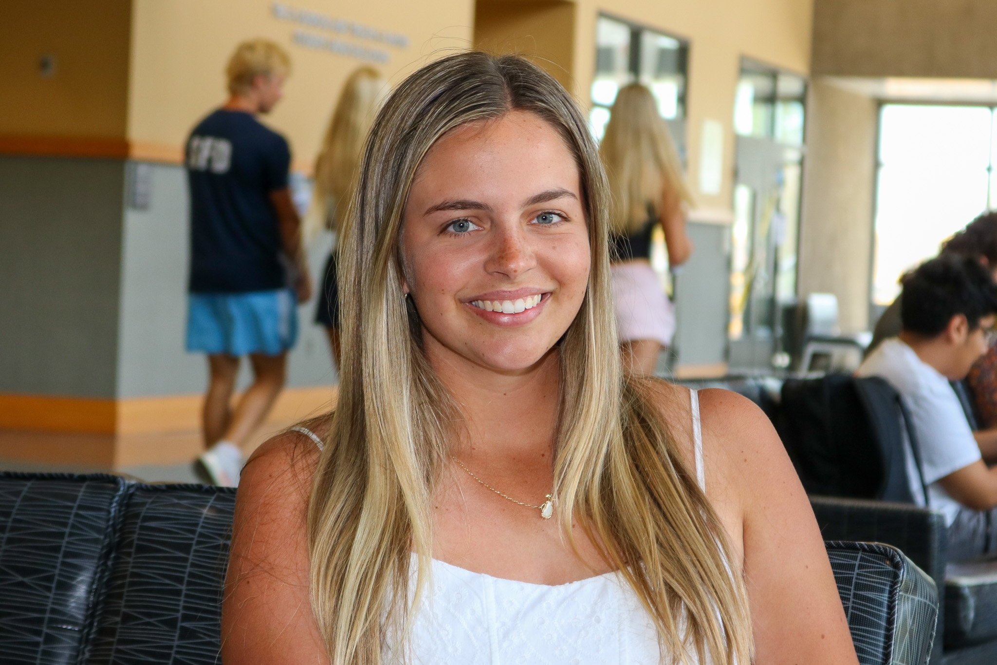Ella Hardy smiles while seated indoors in a campus common area. She has long blonde hair and is wearing a white top and a necklace, with other students and seating visible in the background.